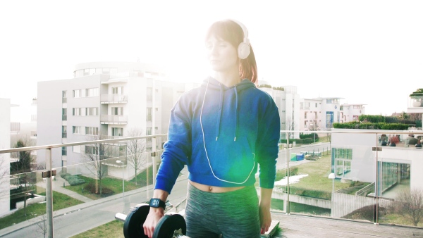 Beautiful young fitness woman with headphones doing exercise with dumbbells at home. Training on a balcony.