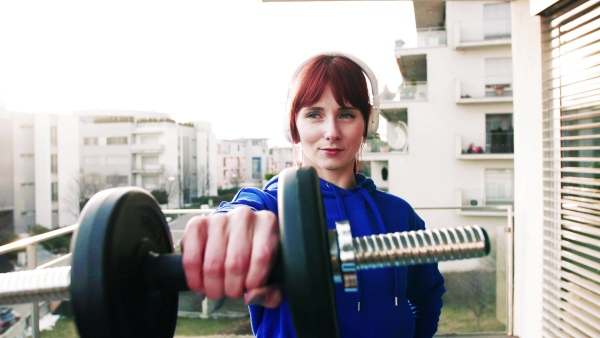 Beautiful young fitness woman with headphones doing exercise with dumbbells at home. Training on a balcony.