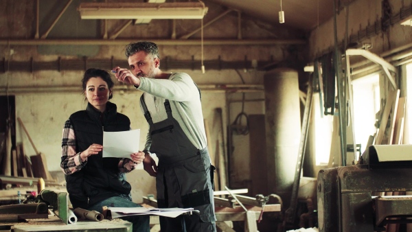 Portrait of a man and woman workers in the carpentry workshop, looking at paper plans.