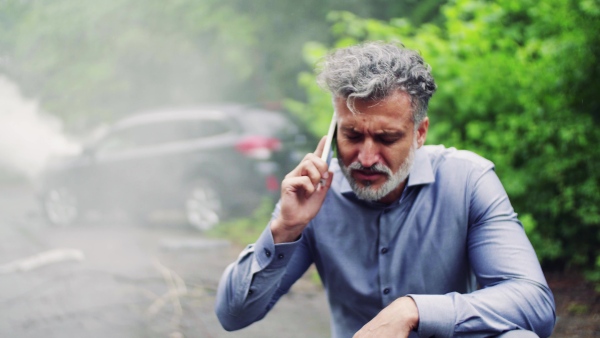 Close-up of a frustrated mature man making a phone call after a car accident, smoke in the background.