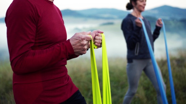 Unrecognizable senior couple doing exercise with resistance rubber bands on meadow in foggy morning in nature.