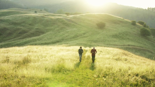 Senior sporty couple running on meadow outdoors in sunny nature at sunrise.