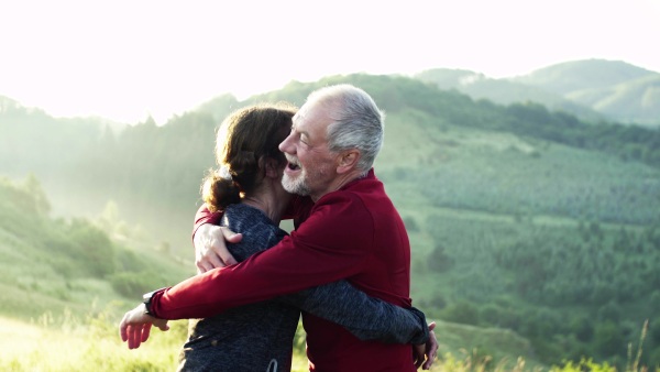 Joyful senior couple runners standing on meadow outdoors at sunrise in nature, hugging.