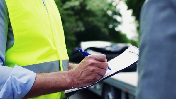 An unrecognizable man insurance agent talking to a woman outside on the road after a car accident, filling in an insurance claim form. A close-up.