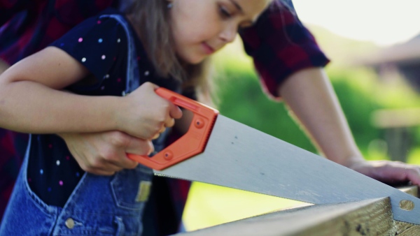 Unrecognizable father and a small daughter with a saw outside, cutting wood.