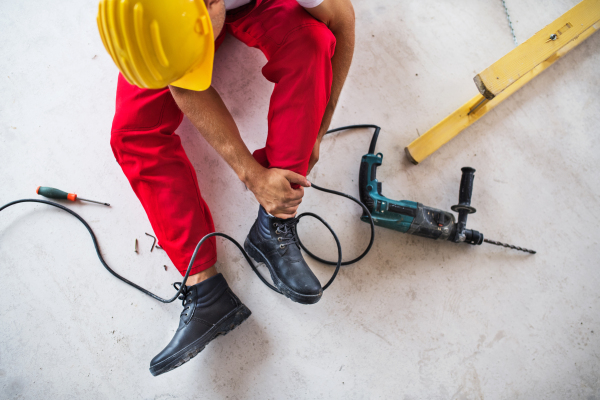 An accident of an unrecognizable male worker at the construction site. A man with an injured leg sitting on the floor. Top view.