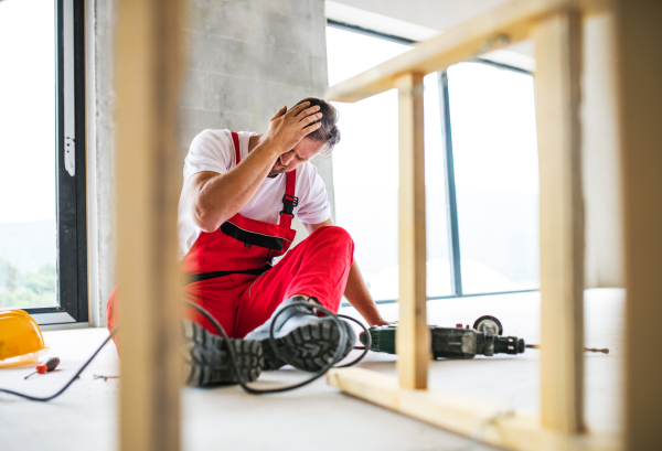 An accident of a male worker at the construction site. A man with an injured head sitting on the floor.