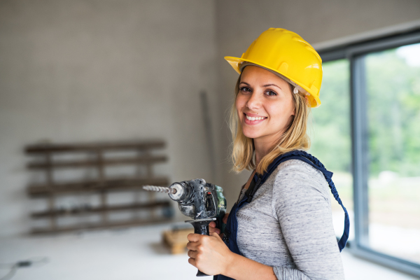 Portrait of a female worker holding an electirc drill on the construction site. Close-up of a beautiful young woman with a yellow helmet. Copy space.