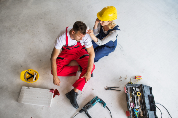 Accident of a male worker at the construction site. A woman with smartphone helping her injured colleague, wiping his forehead. Top view.