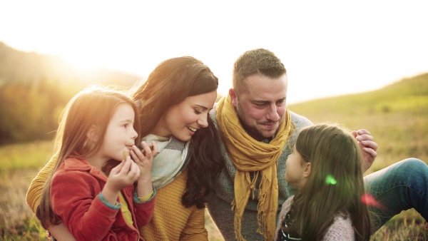 A portrait of happy young family with two small children sitting on a ground in autumn nature at sunset, having picnic. Slow motion.