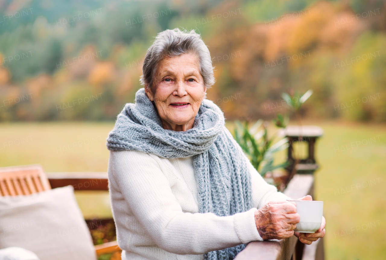 An elderly senior woman with a cup of coffee standing outdoors on a terrace on a sunny day in autumn.