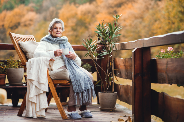 An elderly woman with a cup of tea or coffee sitting outdoors on a terrace on a sunny day in autumn.