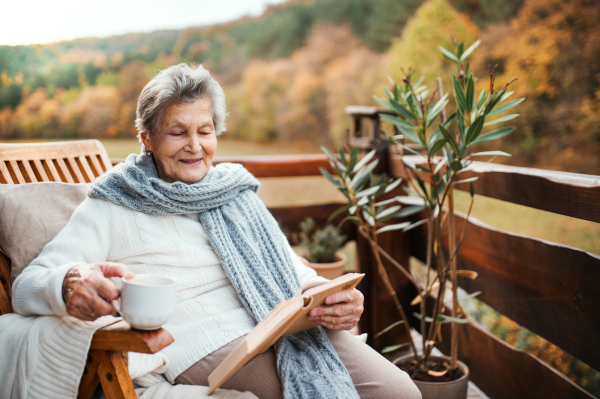 An elderly senior woman with a cup of coffee or tea sitting outdoors on a terrace on a sunny day in autumn, reading a book.