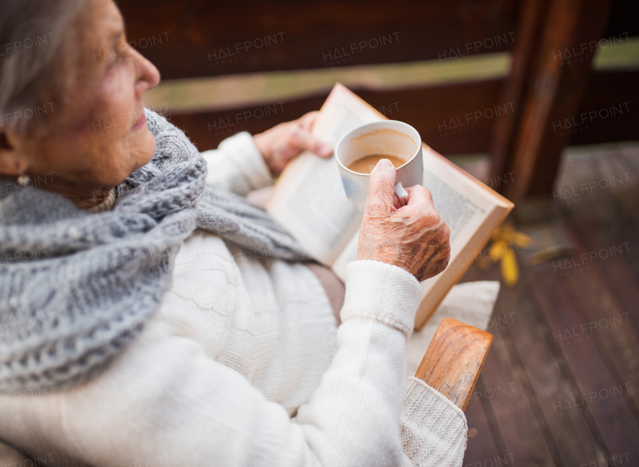 An elderly senior woman with a cup of coffee or tea sitting outdoors on a terrace on a sunny day in autumn, reading a book.