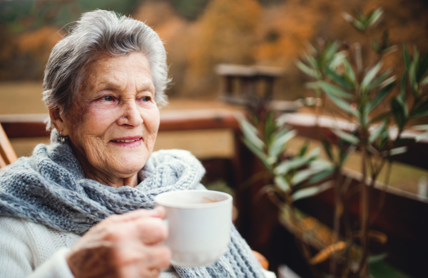 An elderly senior woman standing outdoors on a terrace on a sunny day in autumn.