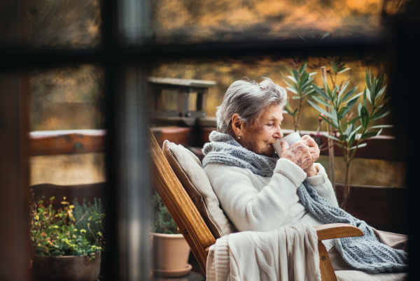 An elderly senior woman sitting outdoors on a terrace in on a sunny day in autumn, drinking coffee. Shot through glass.