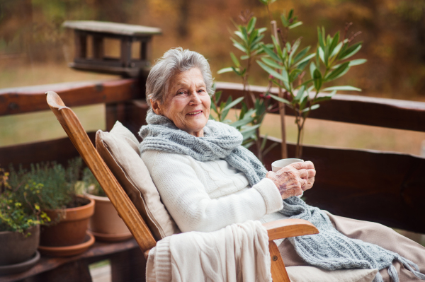 An elderly woman with a cup of tea or coffee sitting outdoors on a terrace on a sunny day in autumn.