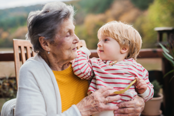 An elderly woman with a toddler great-grandchild sitting outdoors on a terrace on a sunny day in autumn.