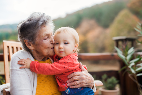 An elderly woman kissing a toddler great-grandchild on a terrace on a sunny day in autumn.