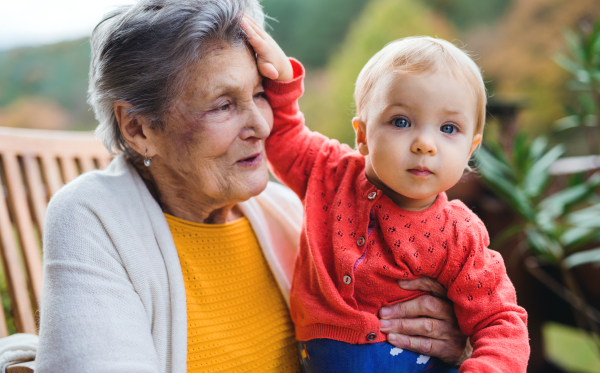 An elderly woman sitting with a toddler great-grandchild on a terrace on a sunny day in autumn.