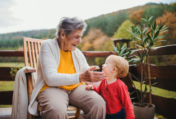 An elderly woman sitting with a toddler great-grandchild on a terrace on a sunny day in autumn.