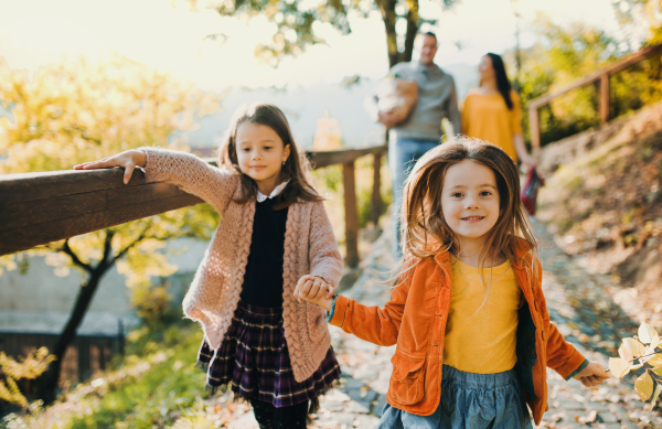 Two girls with unrecognizable parents in the background walking in park in autumn, holding hands.