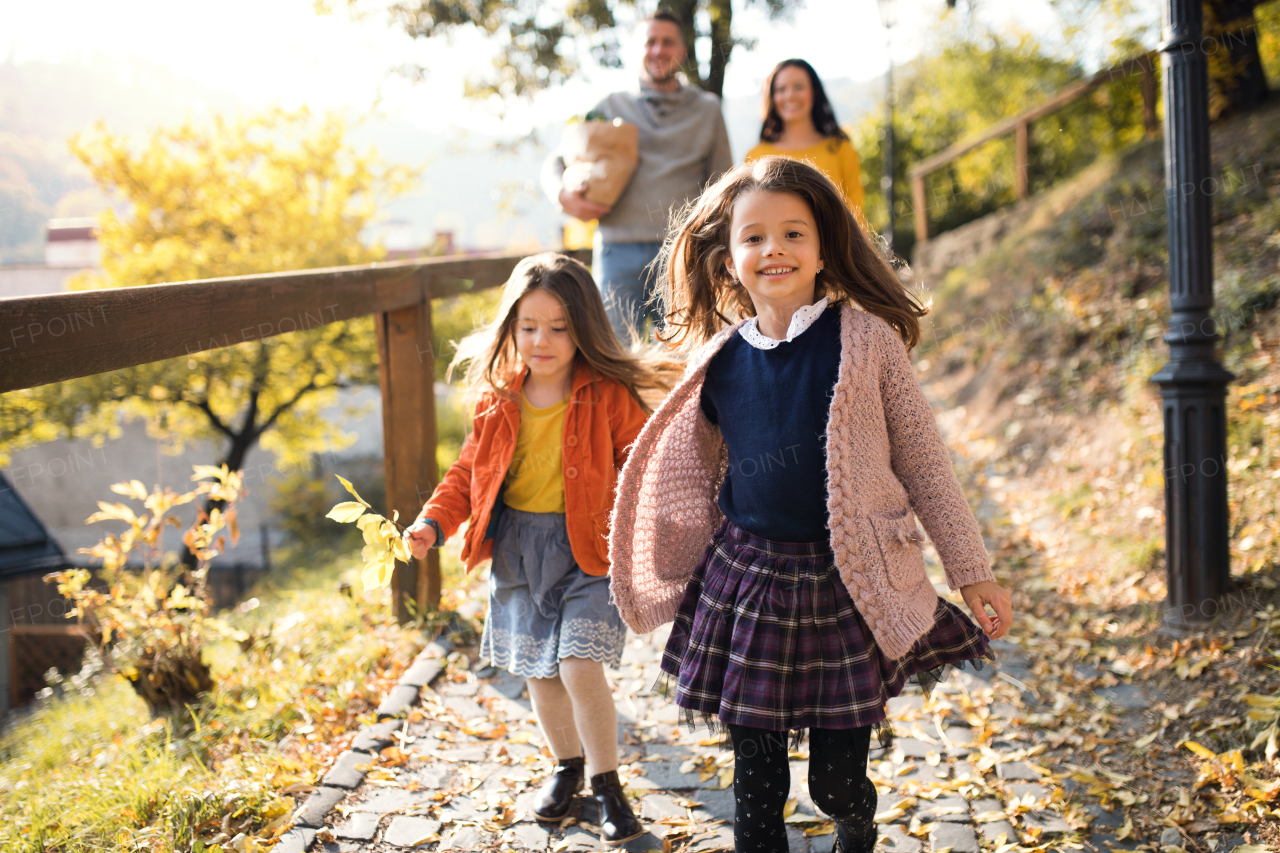 Two girls with unrecognizable parents in the background walking in park in autumn, holding hands.