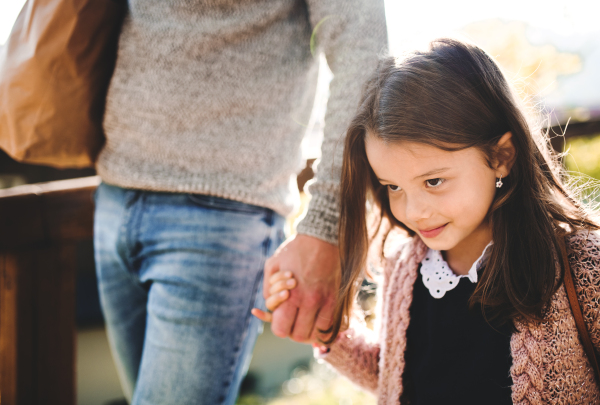 A small girl with unrecognizable father walking outdoors in park in autumn, carrying food in paper bag.