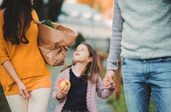 A small girl with apple and unrecognizable parents walking outdoors in park in autumn, carrying food in paper bag.