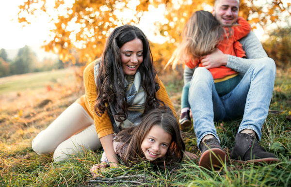 A portrait of happy young family with two small children sitting on a ground in autumn nature at sunset.