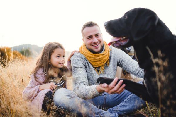 Father with small daughter and a black dog sitting on grass in autumn nature.