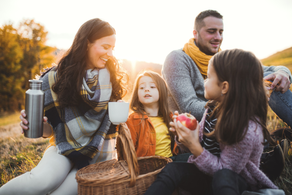 A portrait of happy young family with two small children sitting on a ground in autumn nature at sunset, having picnic.