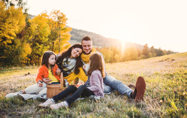 A portrait of happy young family with two small children sitting on a ground in autumn nature at sunset, having picnic.