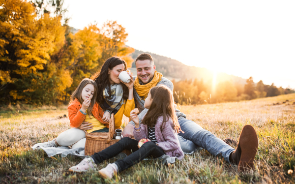 A portrait of happy young family with two small children sitting on a ground in autumn nature at sunset, having picnic.