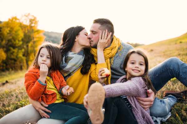 A portrait of happy young family with two small children sitting on a ground in autumn nature at sunset, kissing.