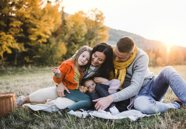 A portrait of happy young family with two small children sitting on a ground in autumn nature at sunset.