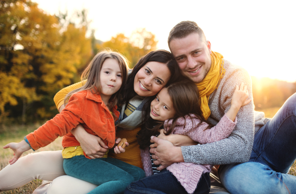 A portrait of happy young family with two small children sitting on a ground in autumn nature at sunset.