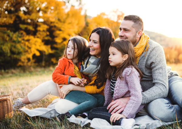 A portrait of happy young family with two small children sitting on a ground in autumn nature at sunset.