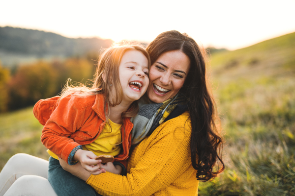 A portrait of young mother with a small daughter sitting on a ground in autumn nature at sunset.