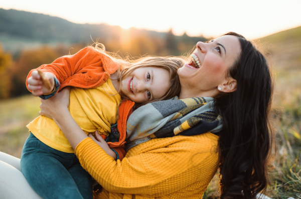 A portrait of young mother with a small daughter sitting on a ground in autumn nature at sunset.