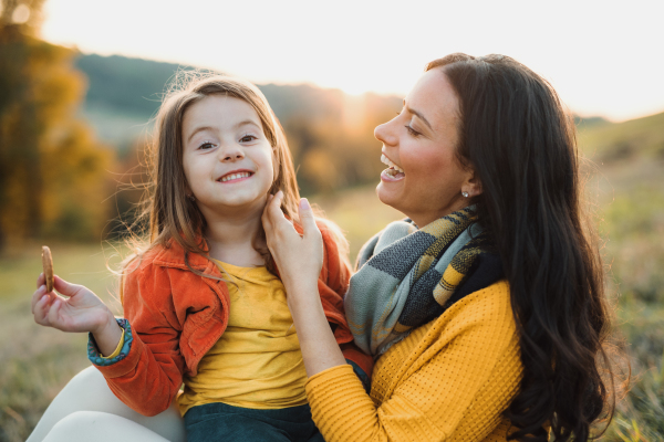 A portrait of young mother with a small daughter sitting on a ground in autumn nature at sunset.