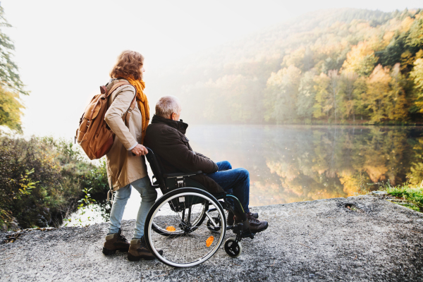 Active senior couple on a walk in a beautiful autumn nature. A woman and man in a wheelchair by the lake in the early morning.