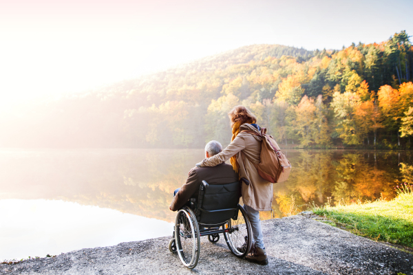 Active senior couple on a walk in a beautiful autumn nature. A woman and man in a wheelchair by the lake in the early morning.