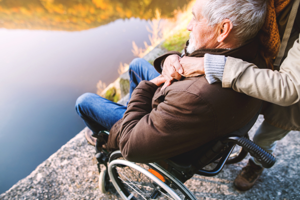 Active senior couple on a walk in a beautiful autumn nature. Unrecognizable woman and man in a wheelchair by the lake in the early morning.