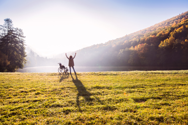 Senior woman standing by the wheelchair in an autumn nature. A woman relaxing by the lake in the early morning. Rear view.