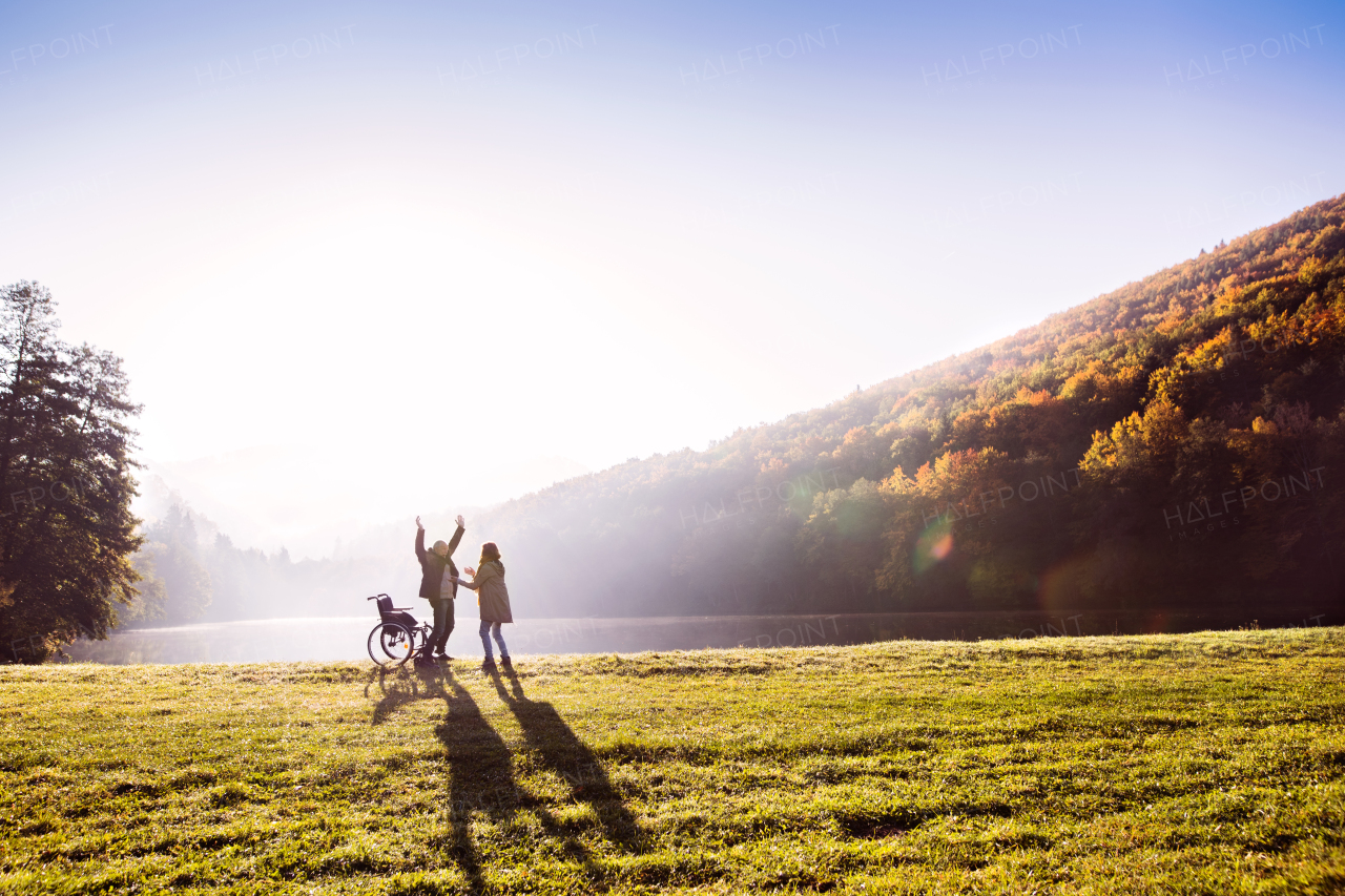 Active senior couple on a walk by the lake in a beautiful autumn nature. A woman and man standing by the wheelchair in the early morning.