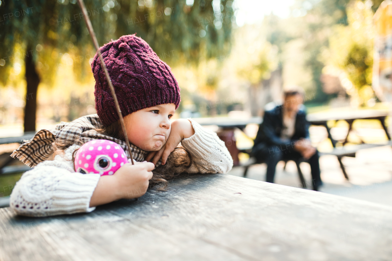 A portrait of a bored small toddler girl sitting in park in autumn nature.