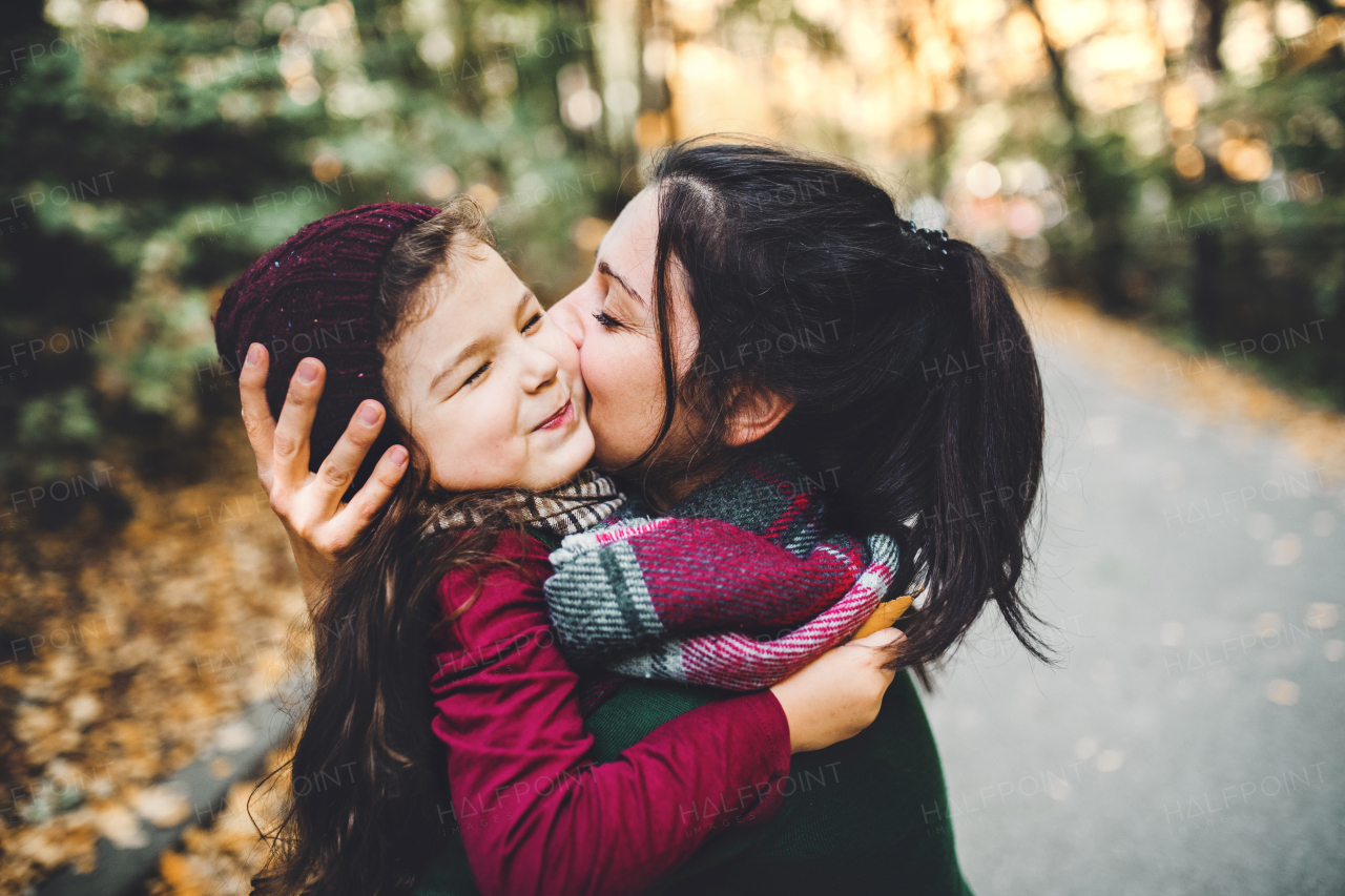 A portrait of young mother with a toddler daughter hugging and kissing in forest in autumn nature.