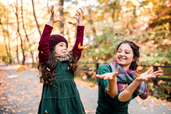 A portrait of cheerful toddler girl with mother in forest in autumn nature, throwing leaves.