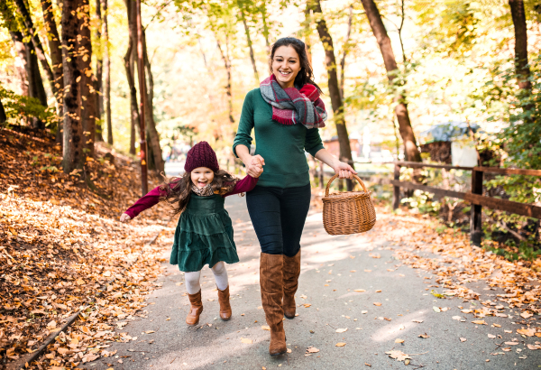 A portrait of young mother with a basket and a toddler daughter running in forest in autumn nature, holding hands.